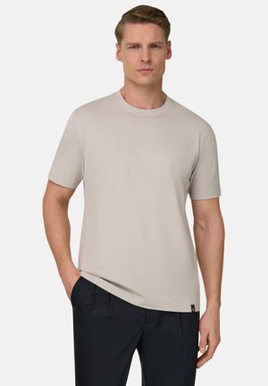 Mężczyzna ubrany w prostą beżową koszulkę na krótkim rękawie z okrągłym dekoltem i czarne spodnie, stojący z jedną ręką w kieszeni na neutralnym tle.