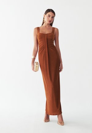Femme portant une longue robe sans manches couleur rouille, tenant une petite pochette beige, avec des boucles d'oreilles, un bracelet doré et des talons ouverts.
