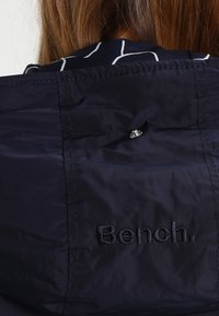 Vue arrière d'une personne portant une veste noire avec un bouton-pression et le nom de la marque brodé "Bench." sur l'arrière du col.