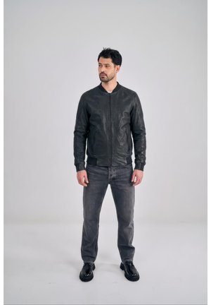 Mann mit kurzen dunklen Haaren und Bart, trägt schwarze Lederjacke, graue Jeans und schwarze Schuhe, steht vor einfachem hellem Hintergrund.