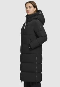 Giacca a vento nera lunga con cappuccio, caratterizzata da un design trapuntato, chiusura con zip frontale, dettagli con logo e tasche laterali. Texture liscia e opaca.