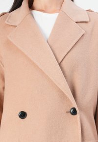 Beige Doppelreiher aus weichem Stoff, mit einem Reverskragen und zwei schwarzen Knöpfen als Akzent. Glatte Textur überall.