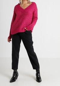 Pull tricoté fuchsia vif avec un col en V profond, une texture côtelée et des manches longues, associé à un jean noir taille haute et des bottines brillantes.