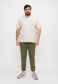 T-shirt beige clair avec un petit logo jaune, associé à un pantalon fuselé vert olive et des baskets grises avec des accents noirs et des semelles blanches.