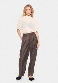 Blusa di tessuto crema con maniche a sbuffo, abbinata a pantaloni marroni a vita alta con pieghe e scarpe nere con punta a stiletto e accenti floreali.