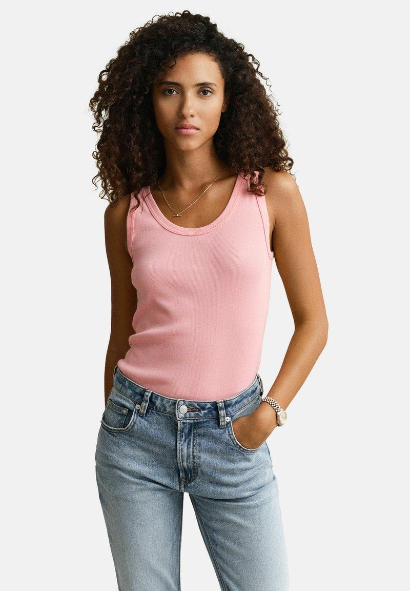 Jeune femme aux cheveux bouclés portant un haut rose sans manches, un jean taille haute bleu clair, une montre et un collier, posant avec une main dans la poche.