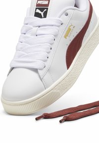 Sneaker bianca Puma Suede XL con striscia marrone, lacci bianchi annodati e un paio extra di lacci marroni accanto alla scarpa.