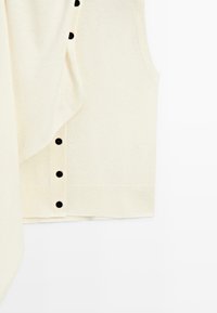 Veste sans manches en tricot couleur crème avec des boutons noirs sur le devant et un ourlet côtelé sur fond blanc.