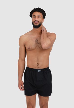 Mann mit Bart und lockigem Haar, der schwarze Boxershorts trägt, mit der linken Hand seinen Hals berührt und leicht nach rechts schaut, vor einem einfachen Hintergrund.