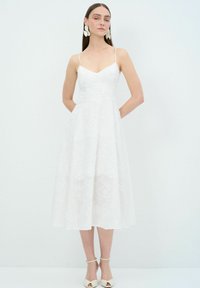 Witte midi-jurk met dunne bandjes, bloemenborduursel en een uitlopende rok. Voorzien van zijzakken en een getailleerde bovenkant, in combinatie met hoge sandalen.