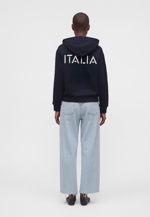 Personne portant un sweat à capuche noir avec "ITALIA" imprimé dans le dos, un jean large bleu clair et des chaussures noires, debout, de dos.