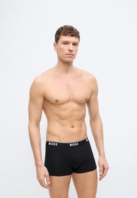 Άντρας χωρίς πουκάμισο, φορώντας μαύρα εσώρουχα BOSS, στέκεται μπροστά σε λευκό φόντο, κοιτάζοντας προς τα εμπρός με ουδέτερη έκφραση.