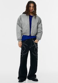 Grijze cropped bomberjack met een zwarte capuchon, gedragen over een blauw overhemd, gecombineerd met zwarte wijde broekspijpen met witte abstracte stiksels.