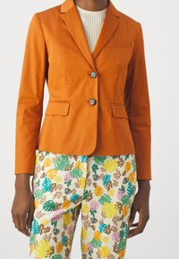 Blazer di cotone arancione con vestibilità su misura, due bottoni e tasche laterali, abbinato a pantaloni a fantasia con foglie multicolori.
