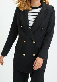 Svart dubbelknäppt kavaj med guldfärgade knappar, slagkrage och strukturerade axlar. Matchas med en randig vit och svart topp under.