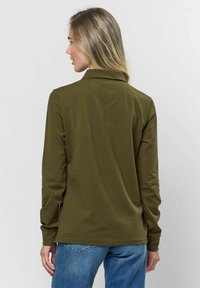 Olajzöld, hosszú ujjú, galléros ing sima textúrával és oldalsó hasítékokkal az alján, világoskék farmernadrággal viselve.