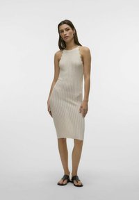 Abito in maglia color beige chiaro, con silhouette aderente, scollo scallop e motivo testurizzato. Abbinato a sandali neri flat.