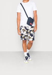Chemise blanche à manches courtes, short bleu marine à motifs de feuilles, sac bandoulière noir avec logo rouge, chaussettes blanches et baskets blanches.