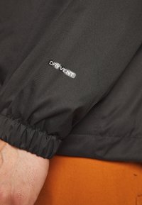 Svart vattentät jackärm med samlad manschett och en "DRYVENT" logo. Slät textur med ett subtilt mönster. Orange bakgrund.