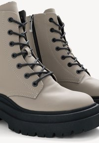 Stivali alla caviglia beige con lacci neri, caratterizzati da una tomaia in pelle texturizzata, suola in gomma e zip laterale per una facile indossabilità.