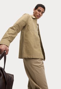 Khaki knopenjack met een zak, gecombineerd met bijpassende broek. Een bruine, gestructureerde leren tas wordt in de hand gehouden.