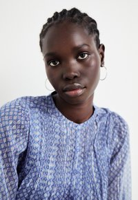 Modra bluzica z vzorcem z gumbom za zapiranje, z rahlim nabiranjem in okroglim izrezom. Ima kratke rokave in lahkoten material.