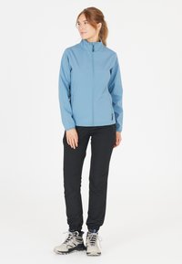 Veste softshell bleue avec un col haut, fermeture éclair frontale et poches latérales ; assortie à un pantalon noir et des chaussures de randonnée de couleur claire.