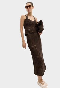 Mujer con top de ganchillo marrón oscuro y falda larga a juego, sosteniendo un gran clutch a juego, con gafas de sol y zapatos blancos tejidos.