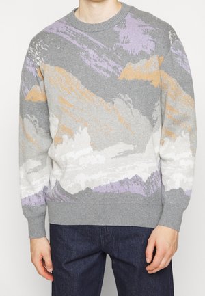 Homme portant un pull en maille gris avec des motifs abstraits orange, blanc et violet, associé à un jean bleu foncé.