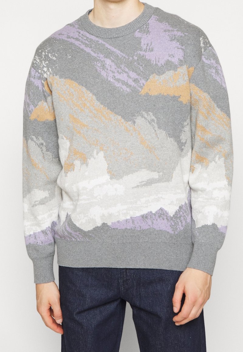 Homme portant un pull en maille gris avec des motifs abstraits orange, blanc et violet, associé à un jean bleu foncé.