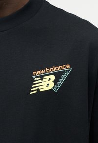 Μαύρο πουκάμισο με πολύχρωμο λογότυπο «new balance NB Running» στο στήθος, ορατό μέρος ενός ατόμου που το φοράει.
