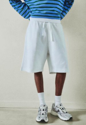 Fehér hálós, zsinóros rövidnadrágot, kék-zöld csíkos hosszú ujjú felsőt, fehér zoknit és szürke sportcipőt viselő férfi beltéren áll.