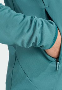 Teal long-sleeve top with a smooth texture, featuring a side zipper and gathered sleeve detail. Airy fabric allows for mobility.