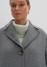 Cappotto in misto lana grigio con colletto a punte, chiusura a un bottone e una dolcevita a coste verde chiaro sotto. Texture liscia.