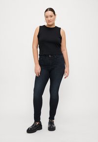 Černý bezrukávový top, tmavě modré úzké džíny a černé platformové boty s kovovými prvky. Modelka stojí s rukama podél těla.