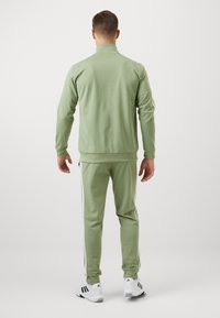 Zeleni trenirka s jaknom na patentni zatvarač i uskim hlačama, s bijelim prugama sa strane. Izrađena od glatkog tkanja s rebrastim manžetama i ovratnikom.