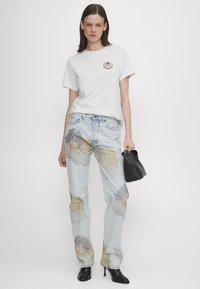 Donna che indossa una t-shirt bianca e jeans azzurri chiari con stampe di volti angelici, tiene in mano una borsa nera e indossa stivaletti neri con il tacco.