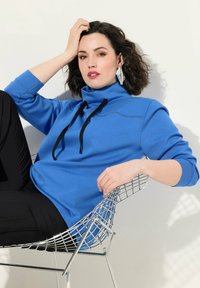 Felpa blu a collo alto con lacci neri e dettagli di cucitura, indossata con pantaloni neri, seduta su una sedia di filo contro uno sfondo chiaro.