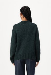 Dunkelgrüner übergroßer Strickpullover mit geripptem Saum und Bündchen, aus strukturiertem Stoff gefertigt, mit Rundhalsausschnitt. Kombiniert mit dunklen Jeans.
