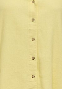 Tissu jaune clair avec une rangée verticale de six petits boutons ronds marron cousus au centre.