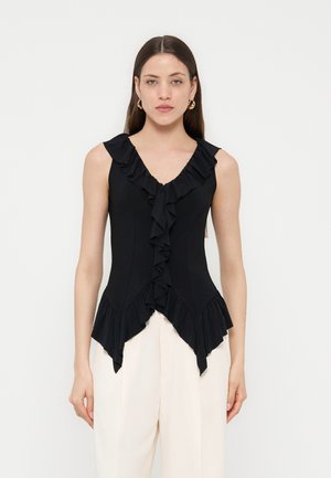 Γυναίκα με μακριά καστανά μαλλιά που φορά χρυσούς κρίκους στα σκουλαρίκια, μαύρη αμάνικη μπλούζα με βολάν και λευκό παντελόνι με ψηλή μέση, στέκεται μπροστά σε λευκό φόντο.