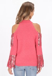 Różowy sweter dzianinowy z wycięciami na ramionach, z frędzlami na rękawach, miękka tekstura i prążkowany dekolt. Noszony z jasnoniebieskimi dżinsami.