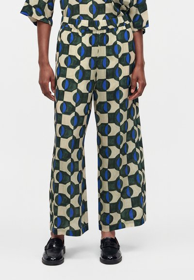 Personne portant un pantalon à jambes larges avec une taille élastique, présentant un motif géométrique de formes bleues et vert foncé sur fond beige, associé à des chaussures noires.