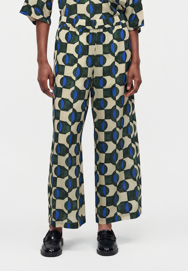 Personne portant un pantalon à jambes larges avec une taille élastique, présentant un motif géométrique de formes bleues et vert foncé sur fond beige, associé à des chaussures noires.