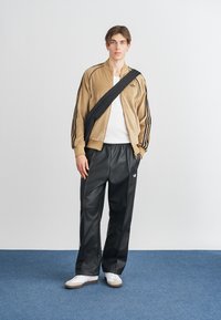 Hnědá sametová bunda na zip s třemi černými pruhy, černé syntetické kalhoty a bílé tenisky s hnědými akcenty. Černá crossbody taška.