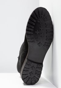 Semelle de botte noire avec des crampons profonds et des motifs en forme de croix, taille 37 indiquée, inclinée contre des murs d'angle blancs.