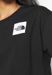 Černé bavlněné tričko s bílým čtvercem, na kterém je v černé barvě vytištěn text a logo "THE NORTH FACE", umístěné na hladkém povrchu.