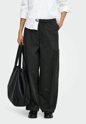 Personne portant un pantalon noir ample avec taille à cordon, chemise blanche, chaussures noires et tenant un grand sac cabas noir dans la main gauche.
