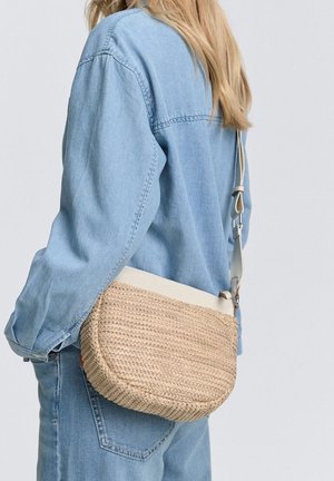 Pletená kabelka přes rameno z slaměných proutků s kulatým tvarem, krémově zbarveným popruhem a jemnou texturou. Doplněná o světle modrý denimový outfit.