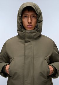 Giacca isolante verde oliva con colletto alto, cappuccio regolabile e grandi tasche frontali. Texture liscia con dettagli delle cuciture e accenti in metallo.
